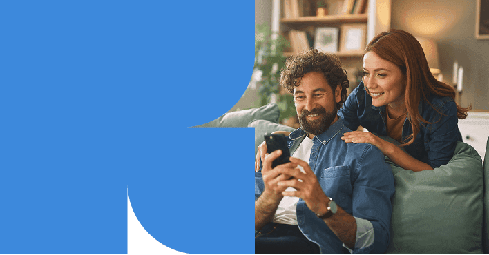 Casal sorrindo enquanto olha o celular no sofá de casa